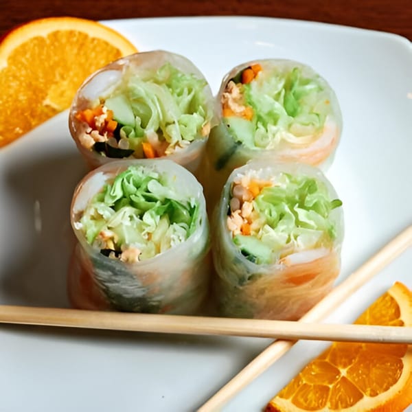 Four fresh spring rolls with lettuce, carrots, and shrimp, served with chopsticks and orange slices.
