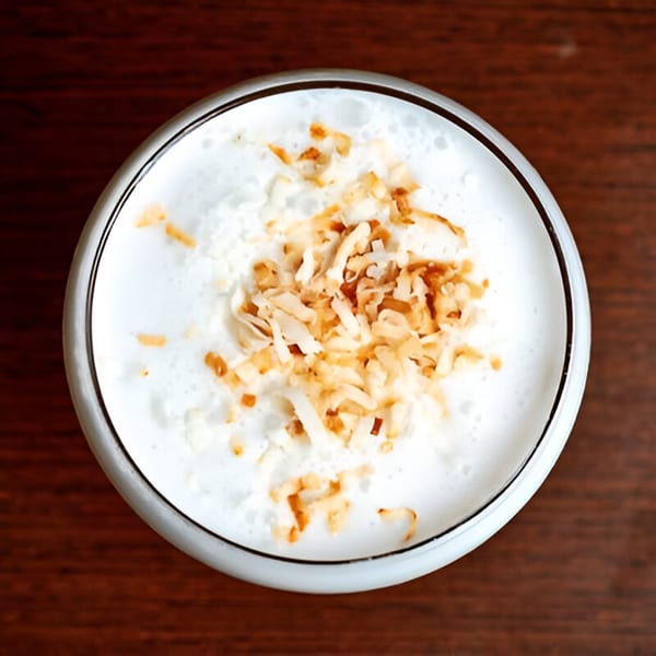A creamy white cocktail with toasted coconut flakes on top, viewed from above on a wooden table.