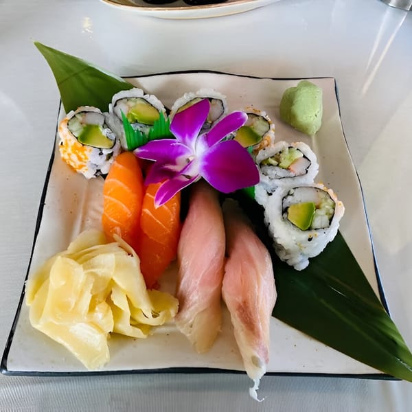 A white rectangular plate of various sushi rolls, sashimi, pickled ginger, and wasabi, garnished with an orchid.